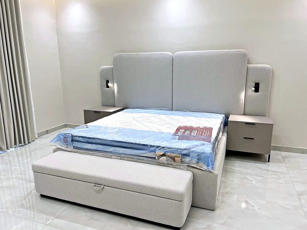 Grey upholstered king bed with double-layer headboard, built-in reading lights and storage ottoman bench by Al Malek Carpentry, Ras Al Khaimah